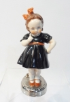 Delicada peça em porcelana alemã em figura de menina com laço com marca na base em rica policromia. Base redonda prateada e vestido negro com laço, sapato coral. Altura de 13 cm e base de 4 cm de diâmetro.